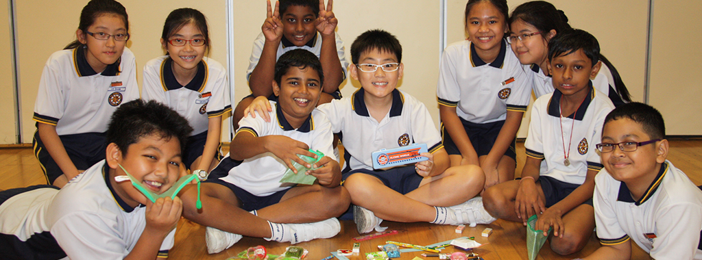 A group of smiling children in white and navy school uniforms sitting on a wooden floor holding paper crafts.